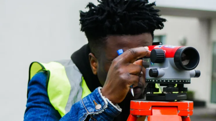 A student looking through a surveyor's telescope, focusing intently on an object through the lens.