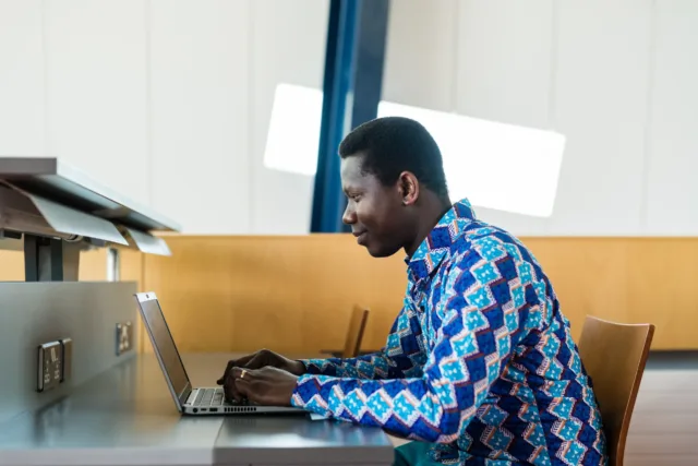 Student in blue patterned shirt sits in library working on his laptop