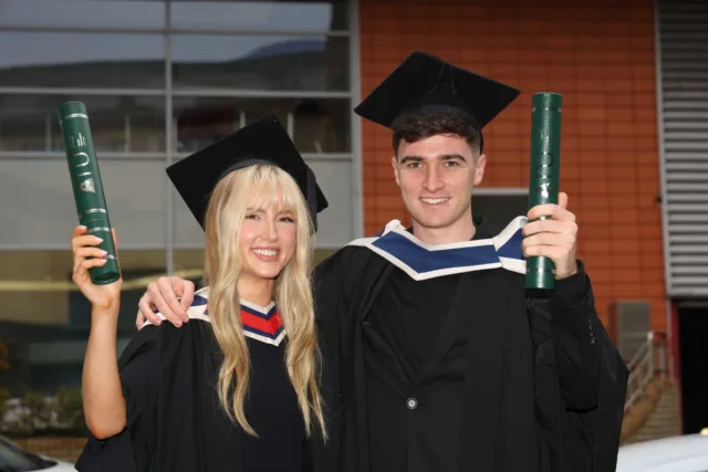 Two students, brother and sister in graduation hat and gown, celebrate their graduation together holding their scrolls in the air