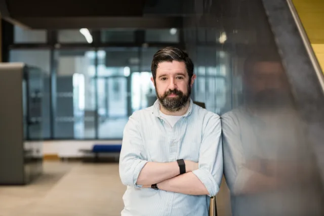 Mature student in white shirt poses with arms folded against wall