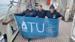 Victoria Matthews, an accessibility activist and ATU Sligo Sport with Business student, alongside her son Luca, an ATU student, and a crew member, holding up the ATU flag onboard a ship