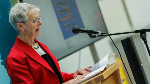 A woman dressed in a red jacket stands at a lectern, reading from the Scrimshaw Journal and speaking into a microphone.