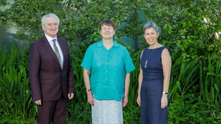 Three people smiling for a photo in front of a green leafy background. The group includes two women and one man, standing closely together.