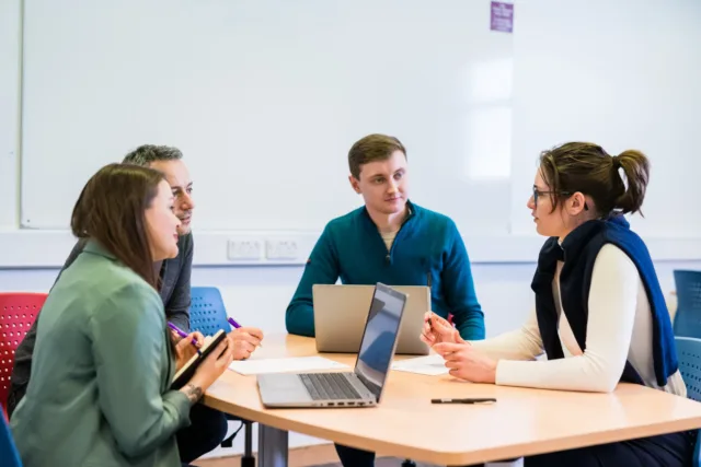 Group of four students, two women and two men, sitting around a table braining storming with laptops