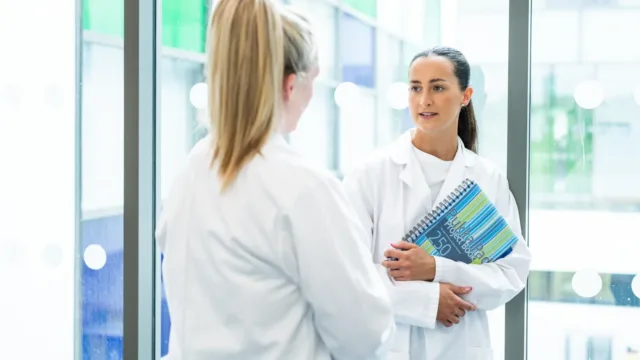 A student in a lab coat holding a notebook faces another student, whose back is to the camera