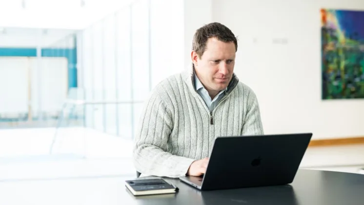 A man typing on a laptop, with a notebook placed on the desk beside him.