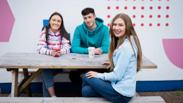 hree students sit on a bench outdoors, smiling and conversing with each other.