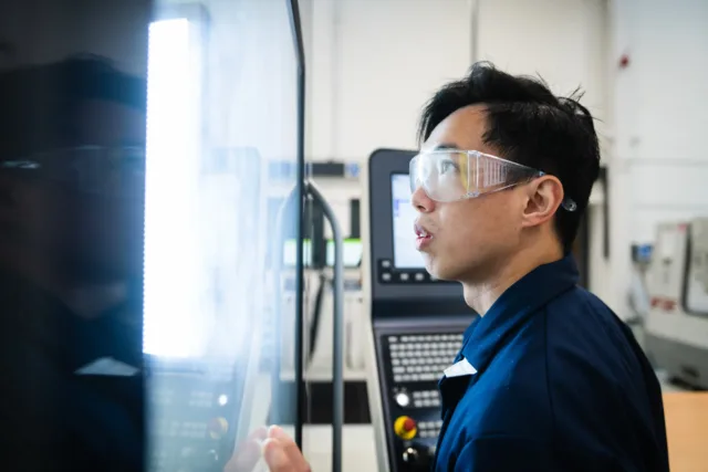 International student in goggles and engineering overalls looking through the glass of a 3D Printer