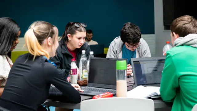 A group of students sitting around a round table, studying. Laptops and water bottles are on the table, and they are focused on their laptop screens.