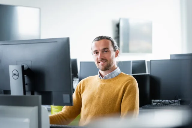 Male student in shirt and yellow jumper sitting at computer in computer lab