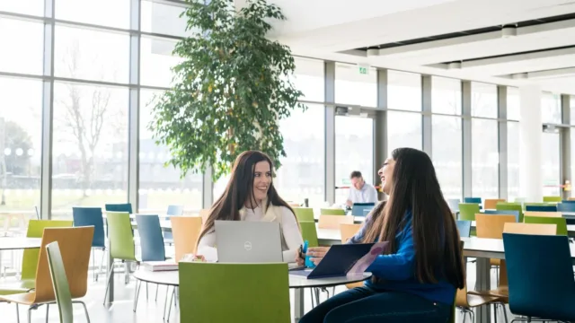 Two women sitting at a table in a bright and airy canteen, with one laptop and a notebook between them. They are smiling and appear happy, engaged in what each other is saying.