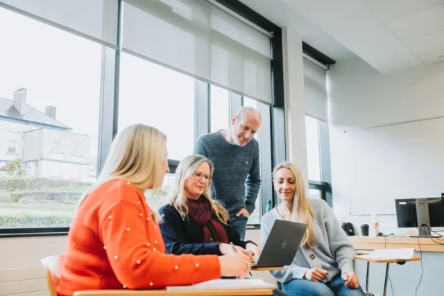 Group of mature students, one male standing behind three females sitting around a laptop.