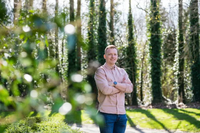 Male student in pink shirt stands arms folded in wooded area.