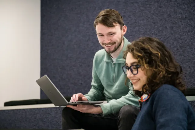 Male and female student laugh while looking at laptop screens