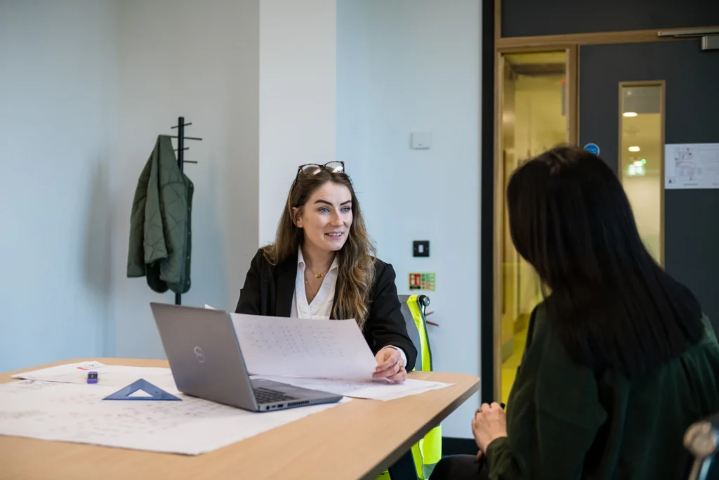 Female worker sits at a chair with high visibility jacket on the back. Laptop open on table showing blueprints to client