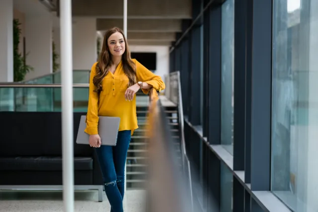 Female student stands i nyellow top with laptop at the top of the stairs in the ATU Mayo Campuse