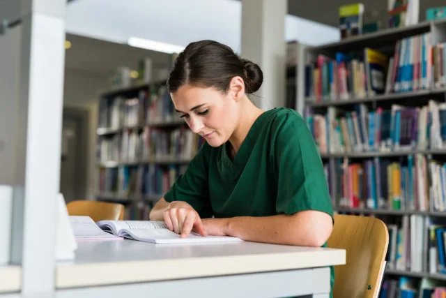 Student nurse in green scrubs reading book in library