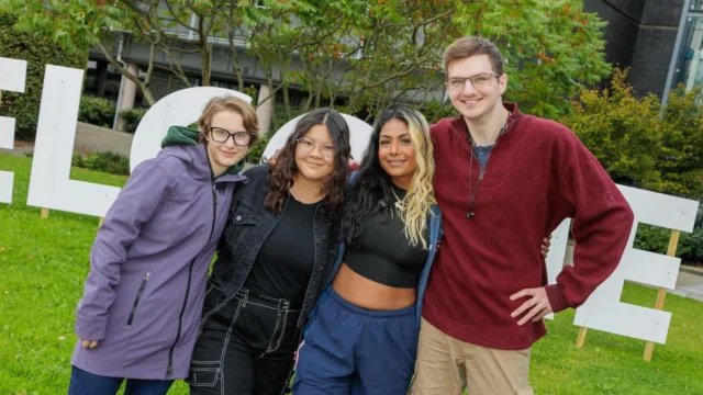 Four students posing in front of a welcome sign, with their arms around one another, smiling for the photo