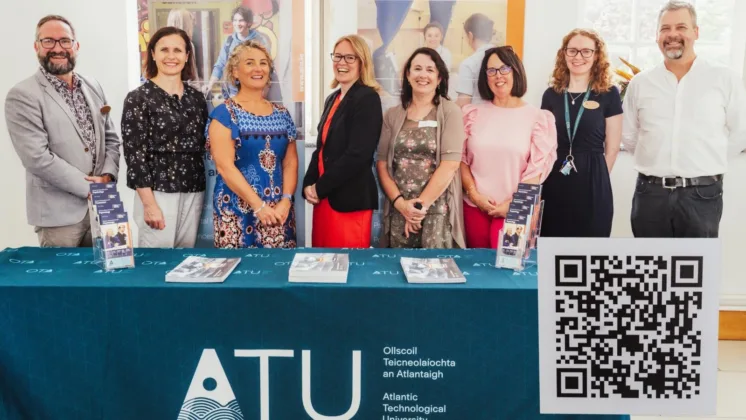 Eight people standing behind a table at an event, smiling at the camera. The table is covered with new books, brochures, and a large QR code attached to the front right. The tablecloth is branded with the ATU logo