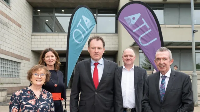 Minister McConalogue stands with ATU staff on the steps outside the main entrance to the Letterkenny campus building. Two ATU teardrop flags are visible in the background. The Minister is visiting the ATU Civil Engineering & Construction Showcase