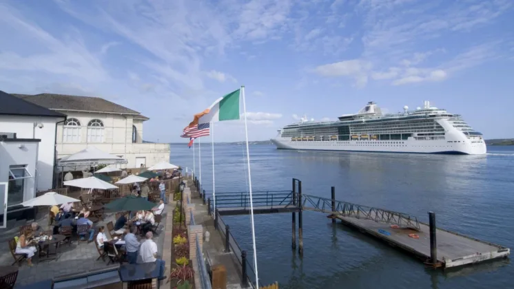 A cruise ship can be seen in the distance, with a pier in the foreground where people are dining. Both the Irish and American flags are flying at full mast on the pier.