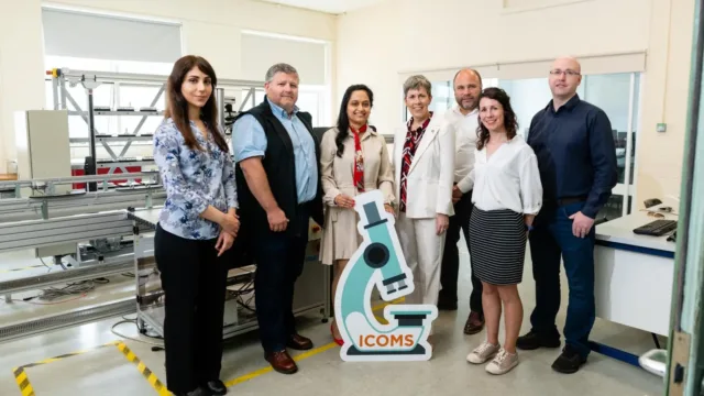 Seven people standing for a photo, smiling at the camera in a lab. In front of them is a sign shaped like a microscope.