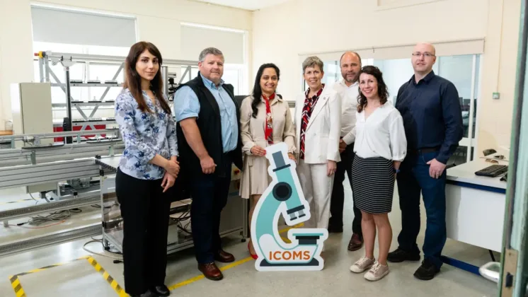 Seven people standing for a photo, smiling at the camera in a lab. In front of them is a sign shaped like a microscope.