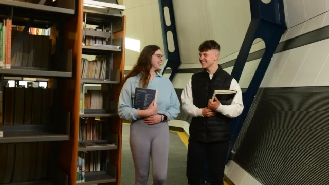 Two students walk together in a library, each holding a book in their hands