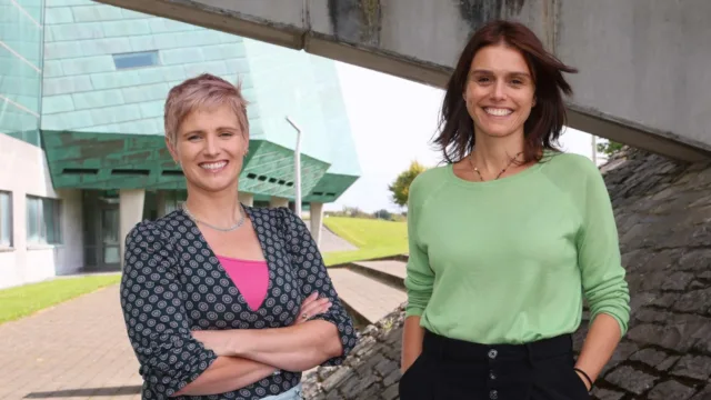 Dr Myra Lydon (University of Galway) with her arms folded and Dr Amaya Vega (ATU) with her arms in her pockets, both smiling towards the camera. The iconic green sails of the Dublin Campus building are visible in the background.