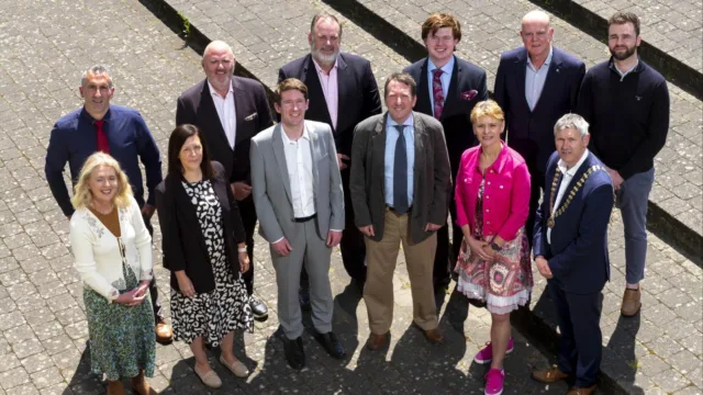 Group of 12 women and men standing on a paved surface, looking up at the camera while squinting in the bright sunlight.