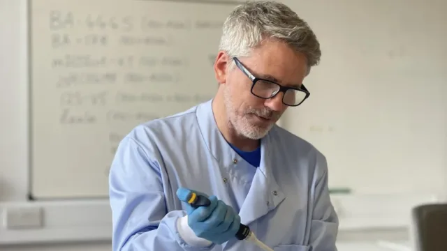 A scientist in a lab, holding a scientific instrument in their hands and wearing blue gloves.