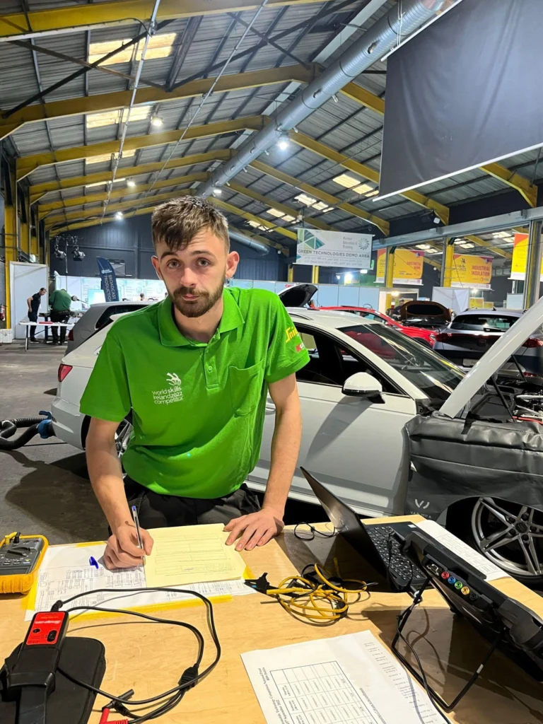 Dion O'Sullivan, ATU student and WorldSkills Ireland Automotive Technology winner, sits at a table writing, with cars in the background.