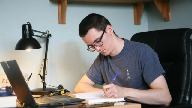 A student studying at their desk, writing in a notebook. A laptop and a lamp are also on the desk.