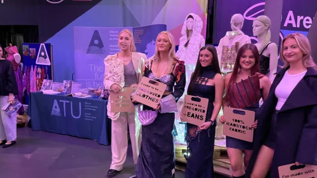 A group of students pose for a photo wearing their Junk Couture outfits, holding up cardboard signs. The students are showcasing creative fashion made from recycled materials