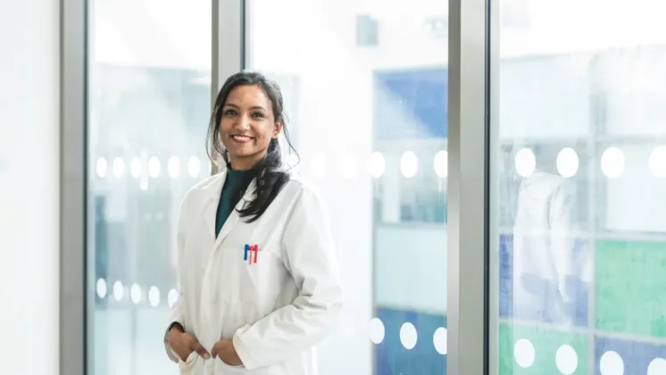 A PhD researcher in a white coat, smiling at the camera with her hands in the pockets of her lab coat. She is standing in a hallway, with a glass window in the background.