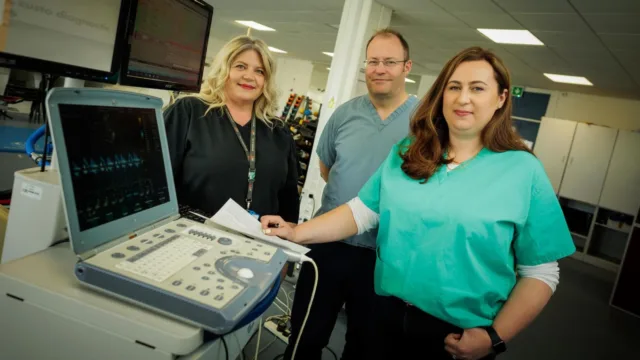 A group of three individuals stand by Clinical Measurement equipment, posing for a photo.