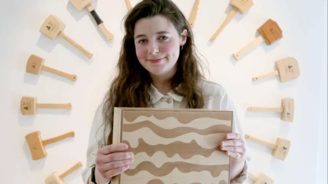 The student, smiling at the camera, holds a wooden box. In the background, wooden mallets are mounted on the wall, arranged like the hands of a clock. The student has long brown hair and wears a light-coloured jacket.