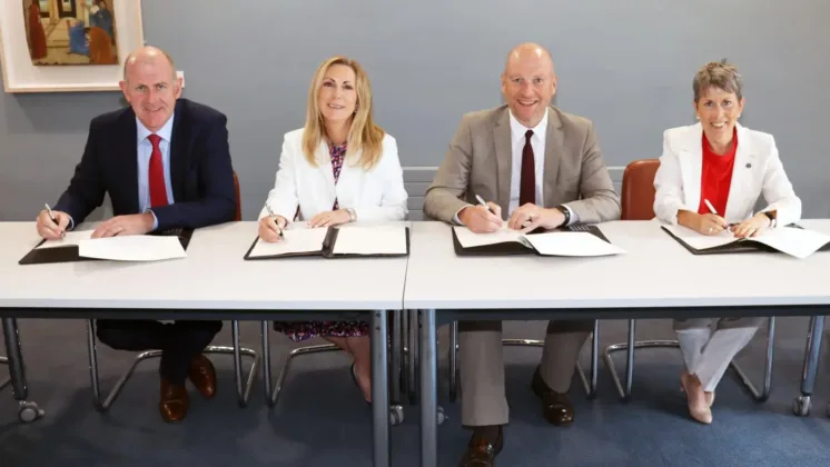 Four people sitting at a table, each holding a pen with a document in front of them, smiling at the camera.