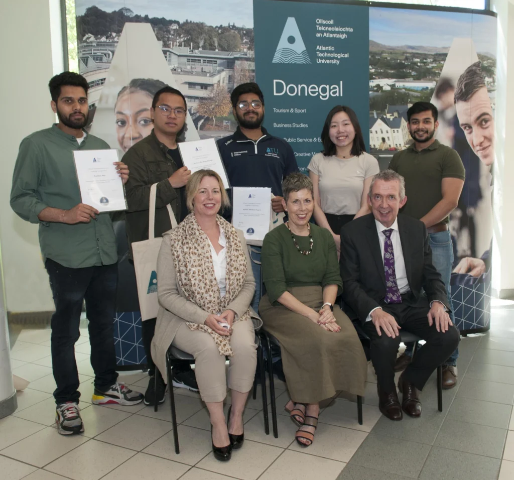 Five students standing at the back, smiling and holding certificates, with three staff members seated in front on chairs, also smiling at the camera.