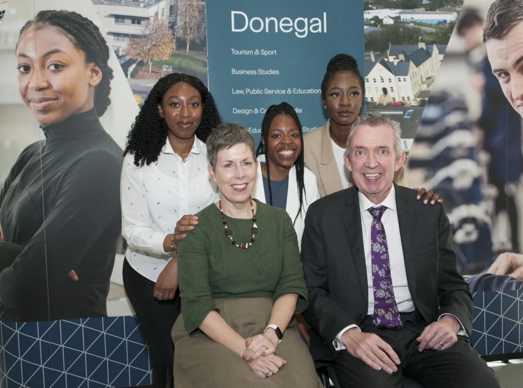 Three women standing at the back, with their hands on the shoulders of the man and woman sitting in front on chairs. All are smiling at the camera.