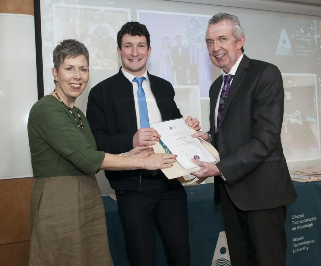 A student is pictured between ATU President Dr Orla Flynn and ATU Donegal Head of College Paul Hannigan. Dr Flynn is presenting an award to the student, who is smiling as they all hold the certificate, facing it towards the camera. Both Dr Flynn and Paul Hannigan stand beside the student, also smiling, with their hands on the certificate.