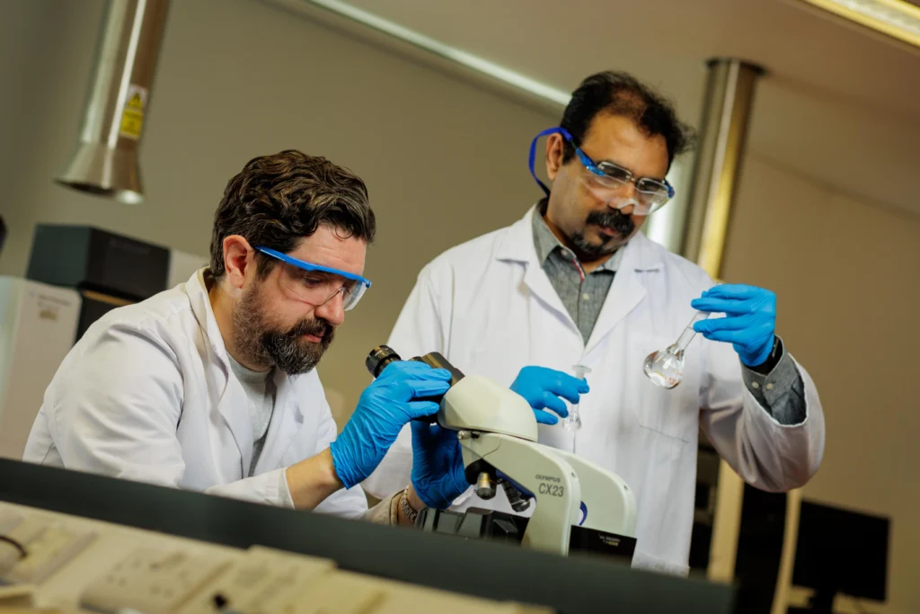 Two men in white lab coats and lab glasses. The man on the left is looking into a microscope, while the man on the right holds two devices containing water, carefully examining them.