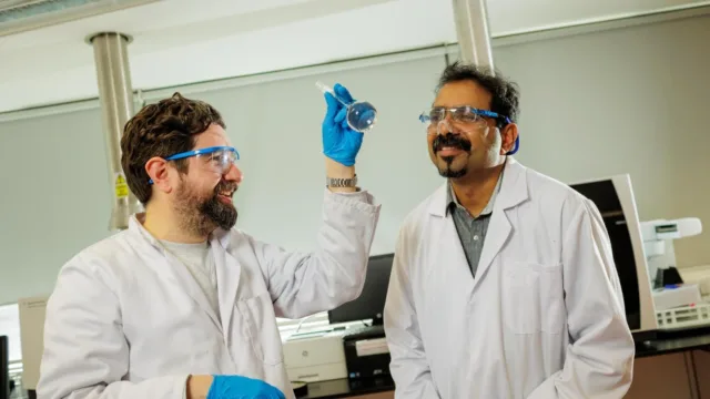 Two men wearing white lab coats, lab glasses, and blue gloves. The man on the left is holding up a device, while both focus on a simple solution aimed at providing clean water.