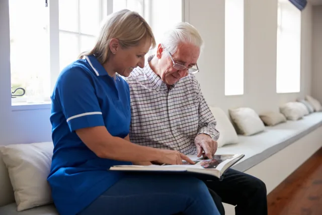 Female nurse in blue scrubs chats with older person while reading a book together