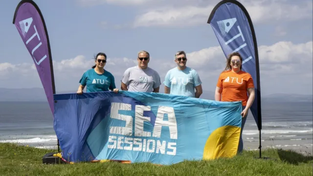 Four people standing between two ATU teardrop flags, holding a banner that reads 'Sea Sessions'. Three of the individuals are wearing brightly coloured ATU tops. The ocean can be seen in the background.