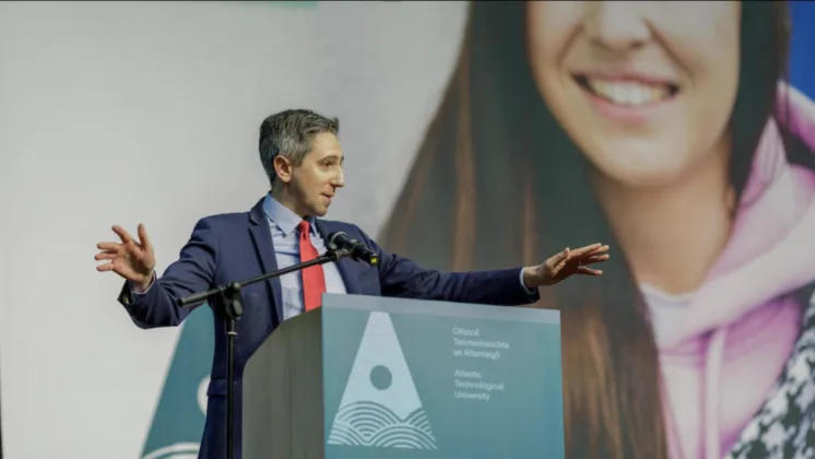 Minister Simon Harris TD speaking from a lectern to a crowd, with his hands extended as he addresses the audience.