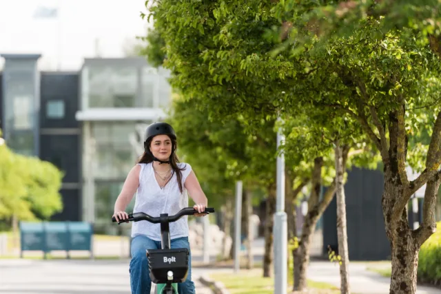 Female student cycling through the ATU Sligo campus on a sunny day with leafy green trees in the background.