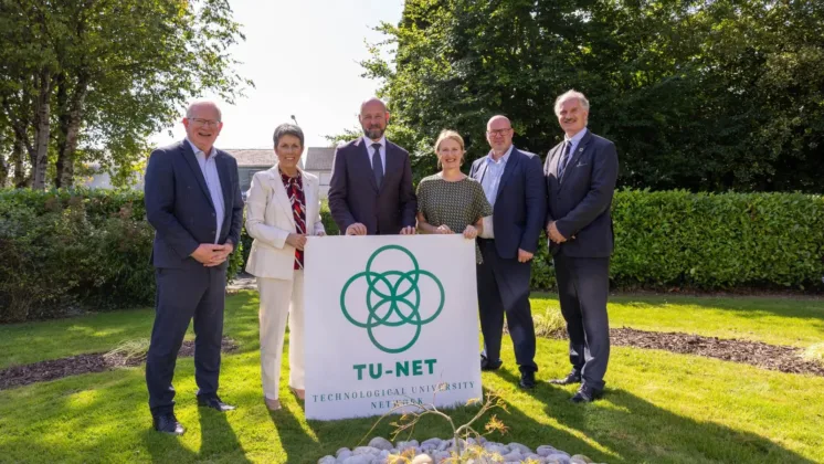 Representatives from the five technological universities (TUs) in Ireland pose for a photo outdoors, holding a sign that reads 'TU NET' (Technological University Network), surrounded by greenery.