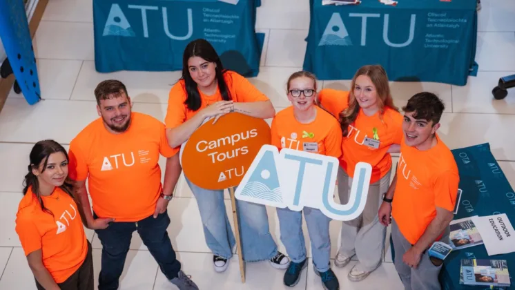 A group of student ambassadors in orange ATU t-shirts, with one holding an ATU sign and another holding a campus tour sign, standing together and looking up for the photo.