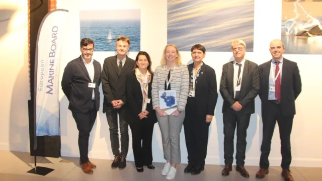 Seven people standing in a row indoors, dressed in business attire, with a banner reading "European Marine Board" and framed ocean-themed photographs on the wall behind them.